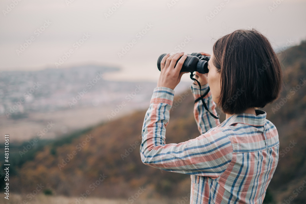 Young woman looks through binoculars and admires the mountain scenery. The traveler has his back turned to the camera.