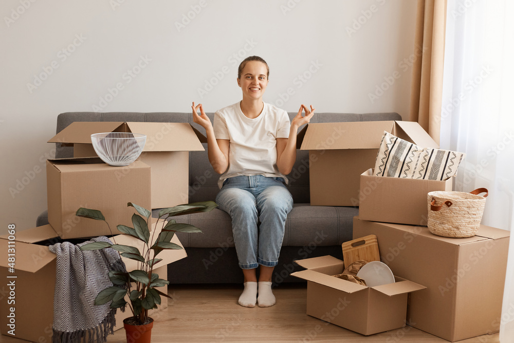 Portrait of smiling satisfied woman wearing white t-shirt and jeans sitting on cough in a new apartment after moving, looking at camera, posing in mudra position, surrounded with belongings in boxes. Portrait of smiling satisfied woman wearing white t-shirt and jeans sitting on cough in a new apartment after moving, looking at camera, posing in mudra position, surrounded with belongings in boxes.