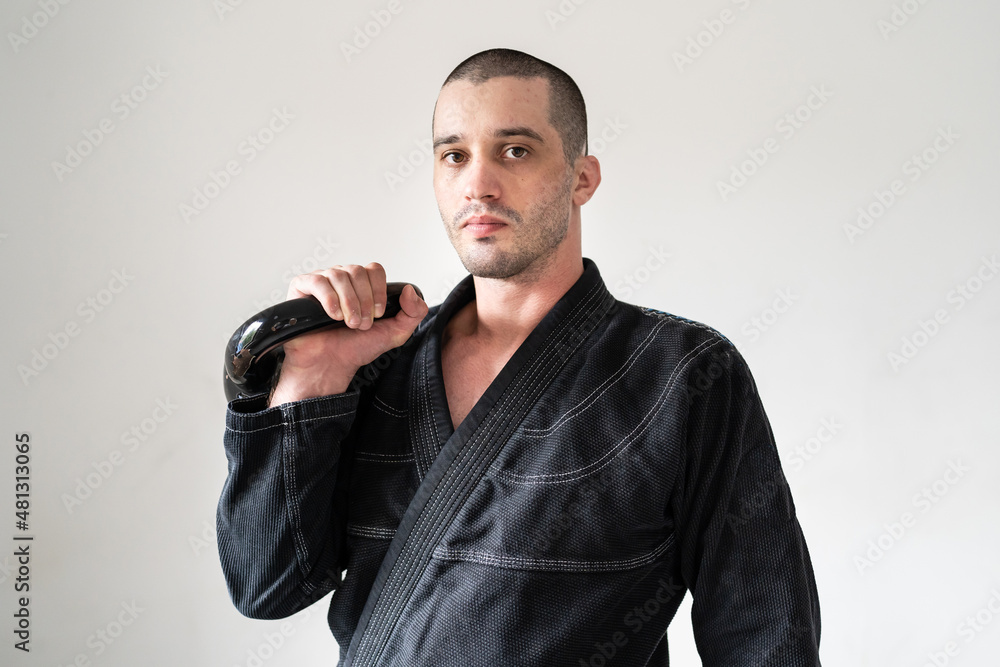 Front view of one man brazilian jiu jitsu bjj athlete standing in front of white wall holding kettlebell weight wearing black kimono gi strength and endurance training concept for martial arts Front view of one man brazilian jiu jitsu bjj athlete standing in front of white wall holding kettlebell weight wearing black kimono gi strength and endurance training concept for martial arts