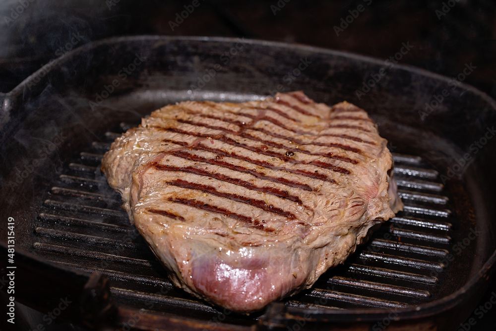 Close-up meat of beef steak grilled in cast-iron ribbed fry pan on gas stove. Delicious juicy meat steak cooking on grill. Close-up meat of beef steak grilled in cast-iron ribbed fry pan on gas stove. Delicious juicy meat steak cooking on grill.