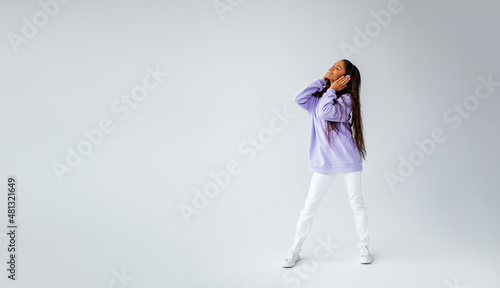 Full length photo of brunette girl with curly hairstyle in purple hoodie and white sneakers and jeans posing on light background. black woman listening to music with headphones. 