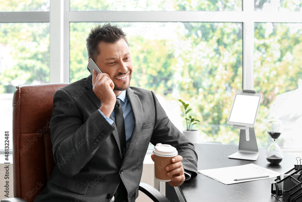 Handsome businessman with cup of coffee talking by mobile phone at table in office Handsome businessman with cup of coffee talking by mobile phone at table in office