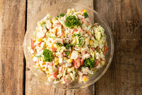 Crab salad with vegetables and crab sticks in a transparent bowl top view. Wooden table.