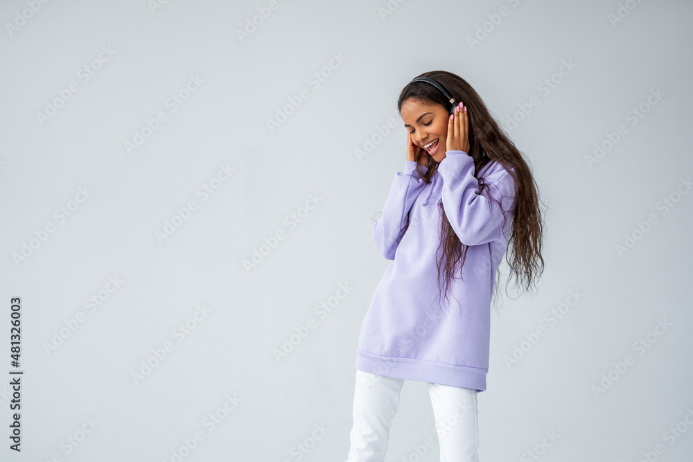 Beautiful brunette girl with curly hairstyle in purple hoodie and white sneakers and jeans posing on light background. black woman listening to music with headphones Beautiful brunette girl with curly hairstyle in purple hoodie and white sneakers and jeans posing on light background. black woman listening to music with headphones
