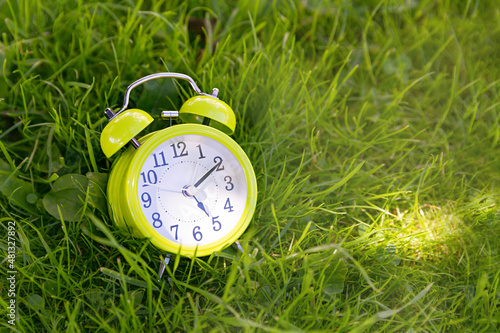 A green alarm clock lies on the green grass, shows 5 o'clock Time for outdoor recreation, a break in work, the end of the working day concept, copy space