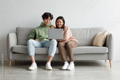 Loving millennial Asian couple watching video on laptop, enjoying spare time, sitting on couch at home, copy space