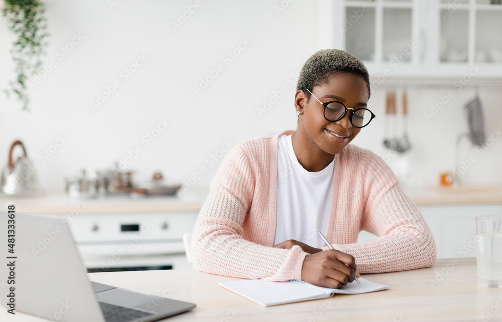 Cheerful millennial pretty black female student or worker in glasses at table with laptop makes notes Cheerful millennial pretty black female student or worker in glasses at table with laptop makes notes