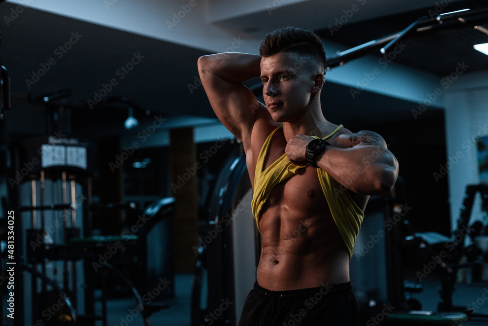 Stylish young sexy sporty man lifted a tank top and shows muscular abdominal, doing a workout in the gym on a dark background Stylish young sexy sporty man lifted a tank top and shows muscular abdominal, doing a workout in the gym on a dark background