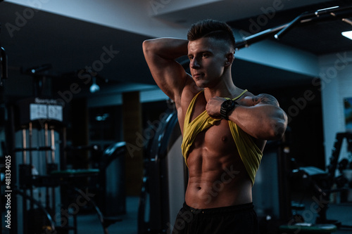 Stylish young sexy sporty man lifted a tank top and shows muscular abdominal, doing a workout in the gym on a dark background