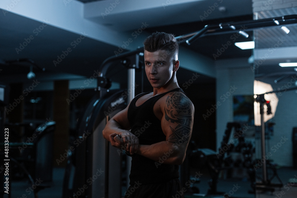 Handsome young sports man with a muscular body with tattoo in a sports black T-shirt with shorts shows muscles in the gym Handsome young sports man with a muscular body with tattoo in a sports black T-shirt with shorts shows muscles in the gym