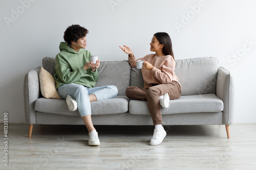 Young Asian boyfriend and girlfriend drinking coffee and chatting while sitting on sofa at home
