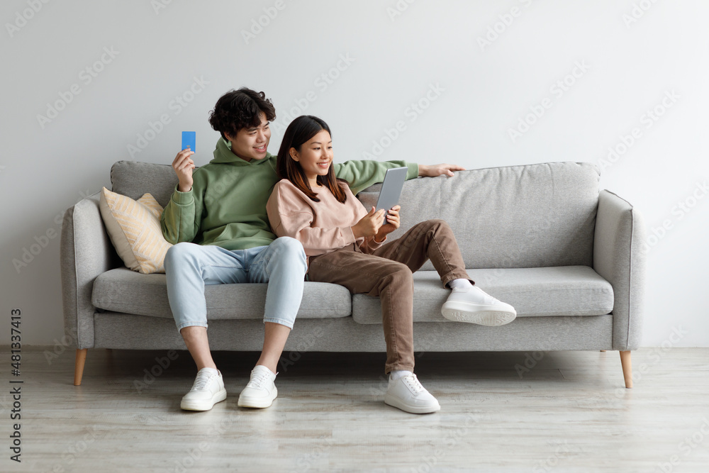 Happy Asian man and woman shopping together on internet, using digital tablet and credit card, sitting on sofa at home Happy Asian man and woman shopping together on internet, using digital tablet and credit card, sitting on sofa at home