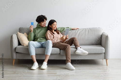 Happy Asian man and woman shopping together on internet, using digital tablet and credit card, sitting on sofa at home