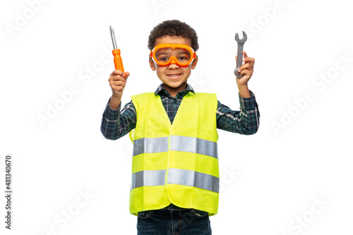 building, construction and profession concept - happy little boy in safety vest and goggles with screwdriver and wrench over white background