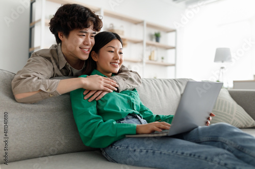 Loving young Asian couple using laptop together, hugging, browsing internet, sitting on couch at home, free space