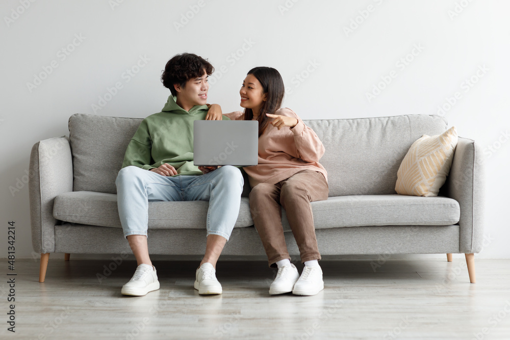 Millennial Asian couple using laptop computer together, sitting on sofa at home, free space Millennial Asian couple using laptop computer together, sitting on sofa at home, free space