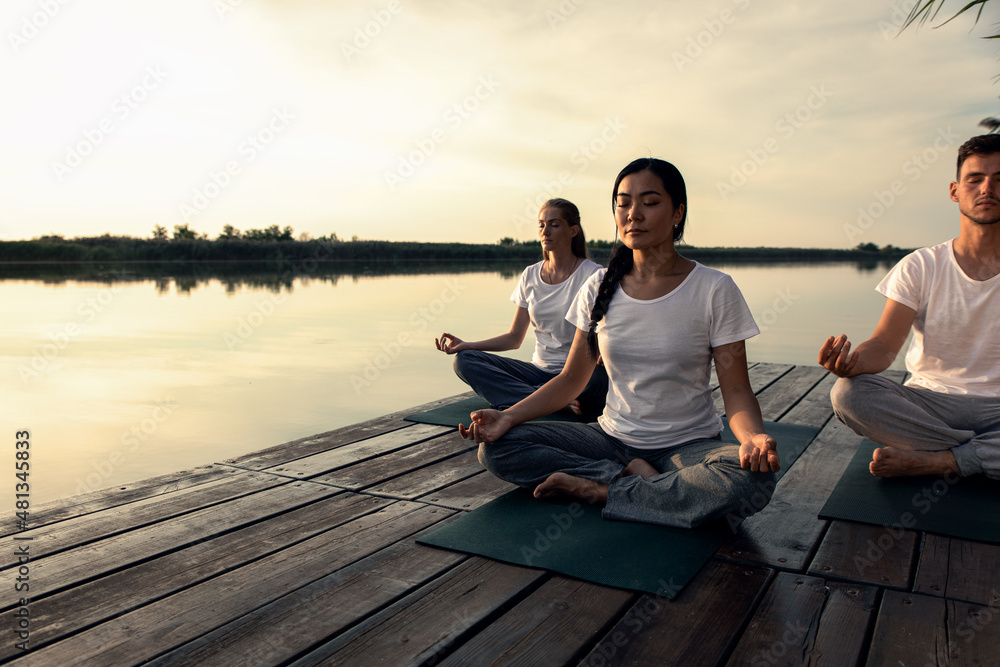 Group of people doing yoga exercises by the lake at sunset.