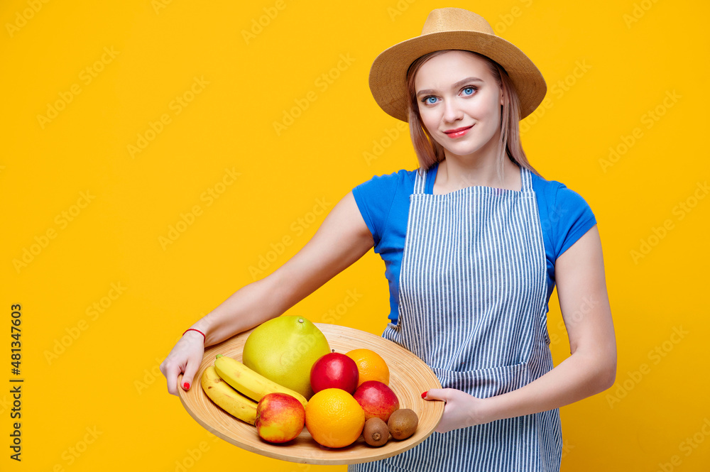 Young female gardener holding fruits banana apple orange kiwi grapefruit in wooden plate dressed in straw hat and an apron on yellow background Young female gardener holding fruits banana apple orange kiwi grapefruit in wooden plate dressed in straw hat and an apron on yellow background
