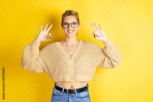 Young beautiful woman wearing casual sweater over isolated yellow background doing ok sign with fingers and smiling, excellent symbol