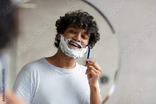 Portrait of indian man shaving near mirror in bathroom, guy with shave foam on face using razor for beauty routine photo