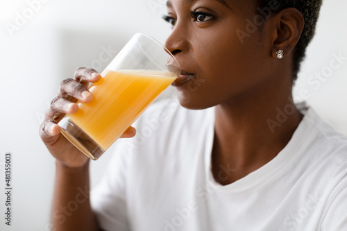 Cropped thirsty young pretty black lady in white t-shirt drinks fresh orange juice from glass indoor, profile