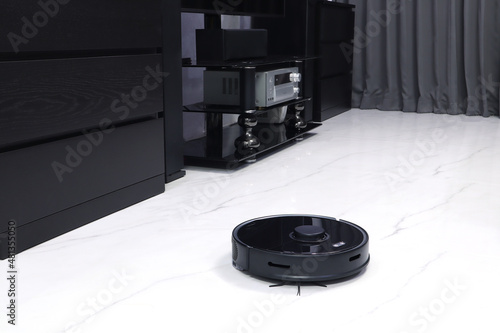 nice black modern automatic scan robotic vacuum is cleaing the dirty marble floor in the nice bedroom with background of cupboard , tv and gray curtain