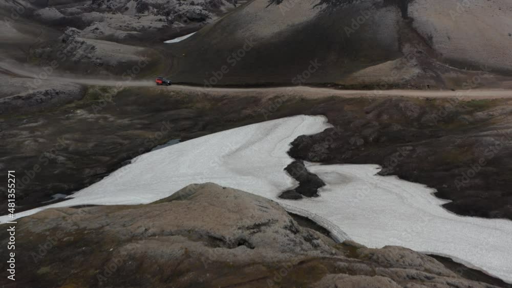 Low flight above volcanic ground. Revealing snowfield and car driving on dirt road in valley. Iceland