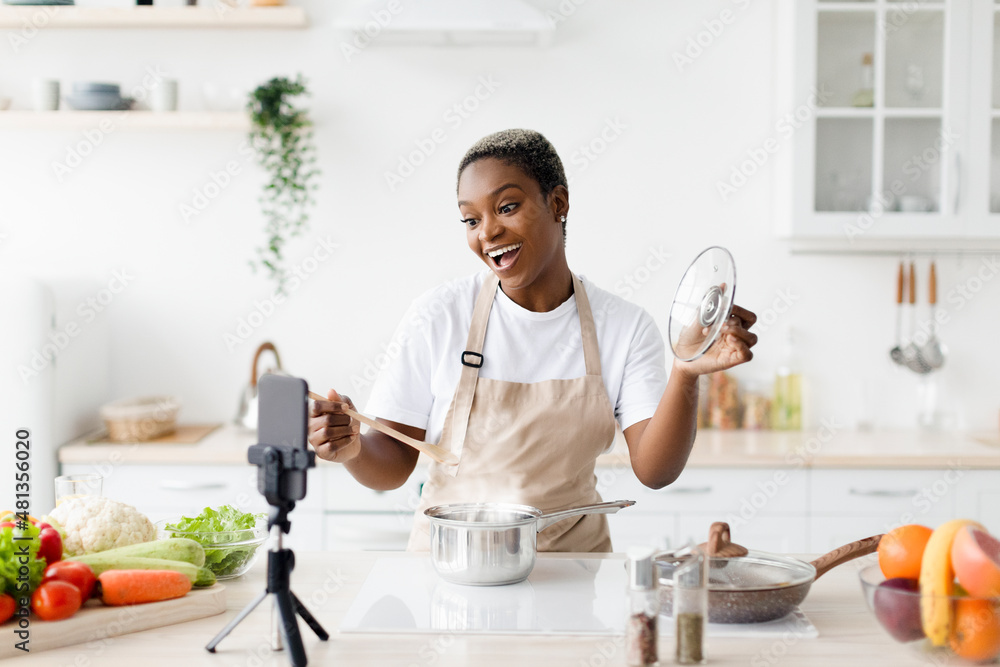 Satisfied millennial black lady with open mouth in apron prepares dinner, shows her dish, shoots video for food blog Satisfied millennial black lady with open mouth in apron prepares dinner, shows her dish, shoots video for food blog
