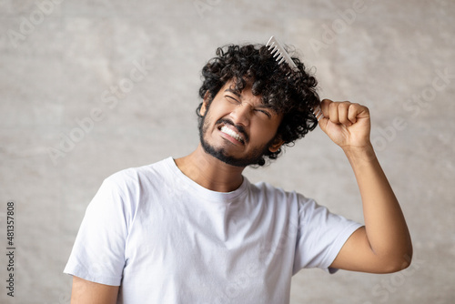 Dry and matted hair. Indian young guy in white t-shirt combining with brush and having pain from tangled curly hair