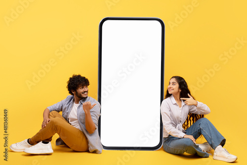 Happy indian couple sitting near big smartphone with blank white screen, demonstrating copy space for app or ad