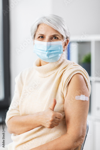 medicine, health and vaccination concept - vaccinated senior woman in mask with medical patch on arm showing thumbs up gesture at hospital