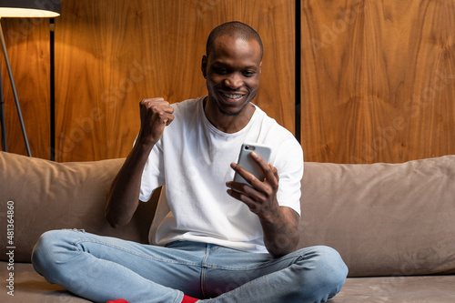 Cheerful black man using a smartphone is sitting at home on the couch, celebrating a victory, winning an online lottery, rejoicing in victory