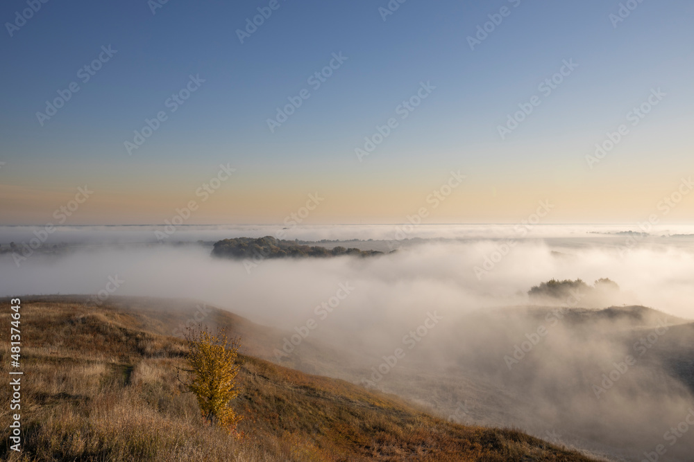 Autumn landscape in the early morning. Fog-covered expanses through which the first rays of the rising sun pass. Trees and hills in the fog. Dawn on a cold autumn morning.