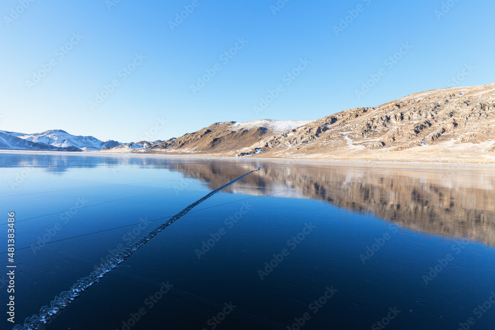 Obraz premium Beautiful winter landscape of frozen Lake Baikal during freezing in the bays. Snow-covered coastal hills are reflected in the expanse of blue ice at sunset. Winter outdoor recreation and ice travel