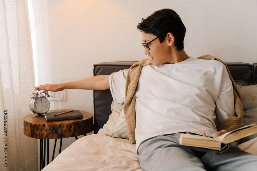 Asian boy in eyeglasses reading book while resting on bed Asian boy in eyeglasses reading book while resting on bed