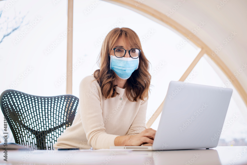 Businesswoman wearing face mask while sitting behind her laptop at office desk Businesswoman wearing face mask while sitting behind her laptop at office desk