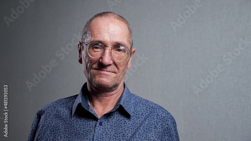 Elderly man with high forehead in classic blue shirt with bad eyesight wears elegant stylish round shaped glasses smiling at grey wall closeup.