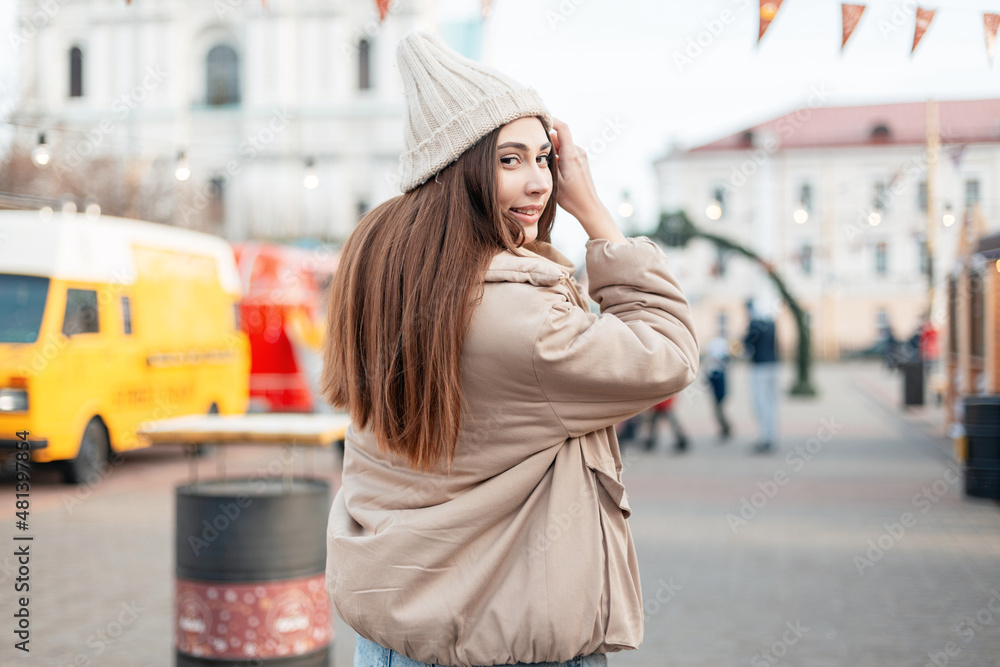 Happy beautiful young smiling woman with long hair in fashionable outerwear with a jacket walks in the city at the fair