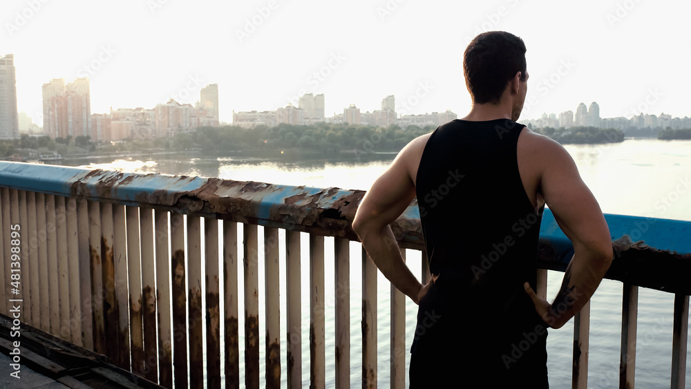 back view of mixed race sportsman standing with hands on hips on bridge over river.