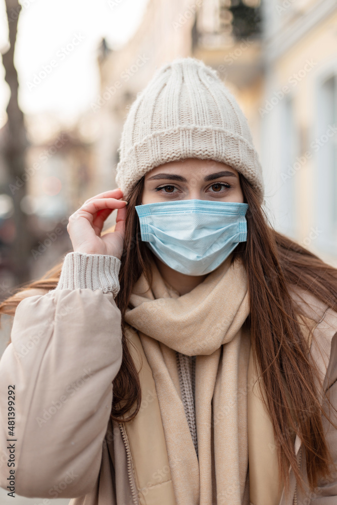 Stylish beautiful young woman with a medical protective mask in fashionable winter clothes with a hat, scarf and jacket walks in the city. Corona virus and pandemic female urban style Stylish beautiful young woman with a medical protective mask in fashionable winter clothes with a hat, scarf and jacket walks in the city. Corona virus and pandemic female urban style