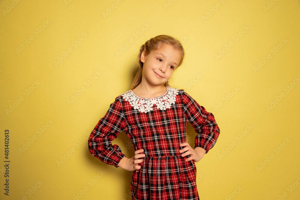 Half-length portrait of cute little girl, pupil wearing plaid dress posing isolated on yellow background. Concept of childhood, emotions, study Half-length portrait of cute little girl, pupil wearing plaid dress posing isolated on yellow background. Concept of childhood, emotions, study