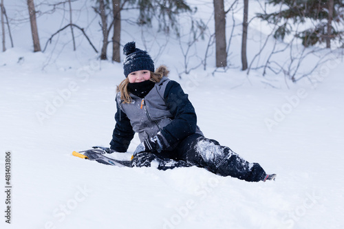 Horizontal portrait of happy pretty little girl in snowsuit sitting down next to sledge after sliding downhill during a winter day
