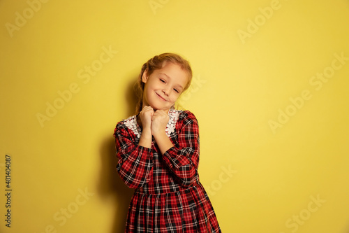 Half-length portrait of cute little girl, pupil wearing plaid dress posing isolated on yellow background. Concept of childhood, emotions, study