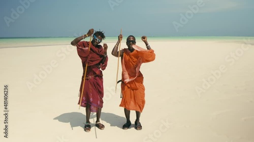 Two Young Happy Masai Tribe Members in Traditional Red Dresses and Sunglasses Dancing on the Sand Beach of the Indian Ocean. Zanzibar. Tanzania. 4K 50fps photo