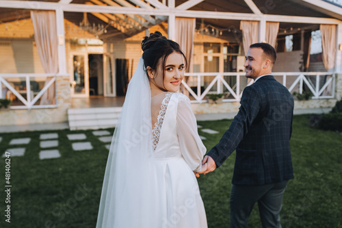 A stylish groom and a beautiful, smiling brunette bride in a white dress are walking along the green grass, in nature, against the backdrop of a cottage, at home. Wedding portrait, photography.