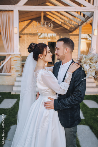 A young groom and a beautiful, smiling brunette bride in a white dress stand embracing on the green grass, in nature, against the backdrop of a cottage, at home. Wedding portrait, photography.