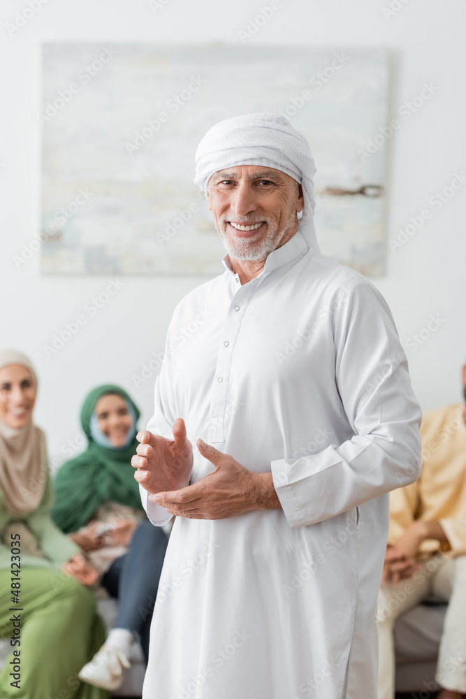 happy muslim man in white turban looking at camera near blurred family. happy muslim man in white turban looking at camera near blurred family.