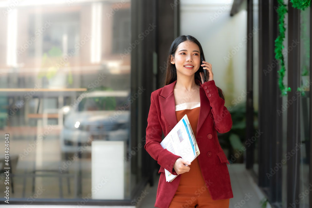 Confident Asian businesswoman in a red suit standing with document talking on the phone with her client outside the office. Confident Asian businesswoman in a red suit standing with document talking on the phone with her client outside the office.
