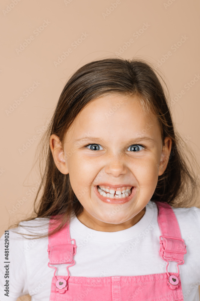 Portrait of female kid with scary, frightening grinning forced smile with all teeth with playful shining eyes looking at camera wearing bright pink jumpsuit and white t-shirt on beige background. Portrait of female kid with scary, frightening grinning forced smile with all teeth with playful shining eyes looking at camera wearing bright pink jumpsuit and white t-shirt on beige background.