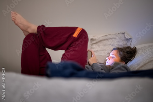 Dark room in evening night or morning and young woman girl lying down on bed with mobile phone smartphone using social media and blue light reflection on face